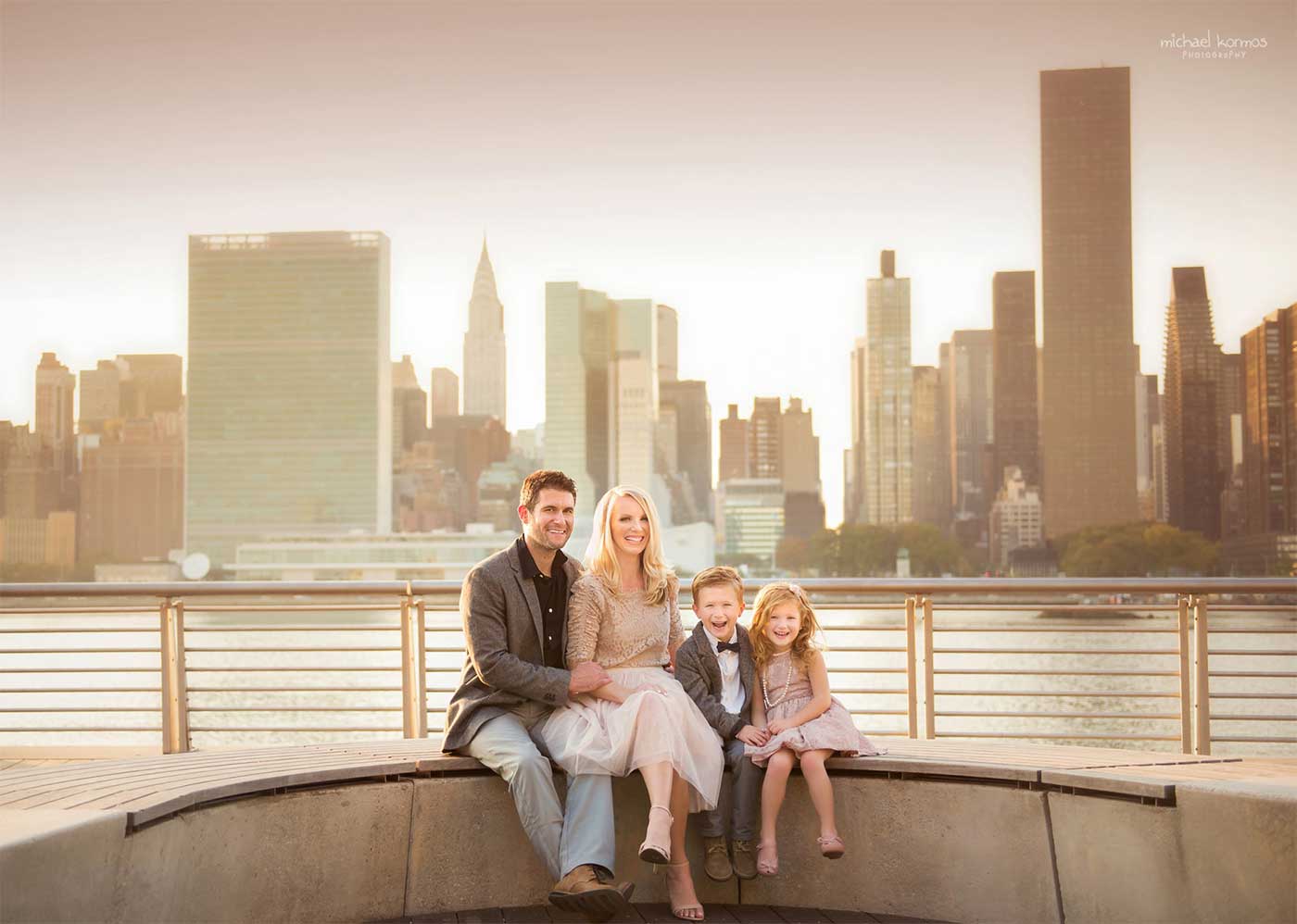 A family of four is sitting on a curved bench by the waterfront with the NYC skyline in the background. The parents are seated close, their two young children smiling between them. The sun is setting, casting a warm glow over this perfect family moment.