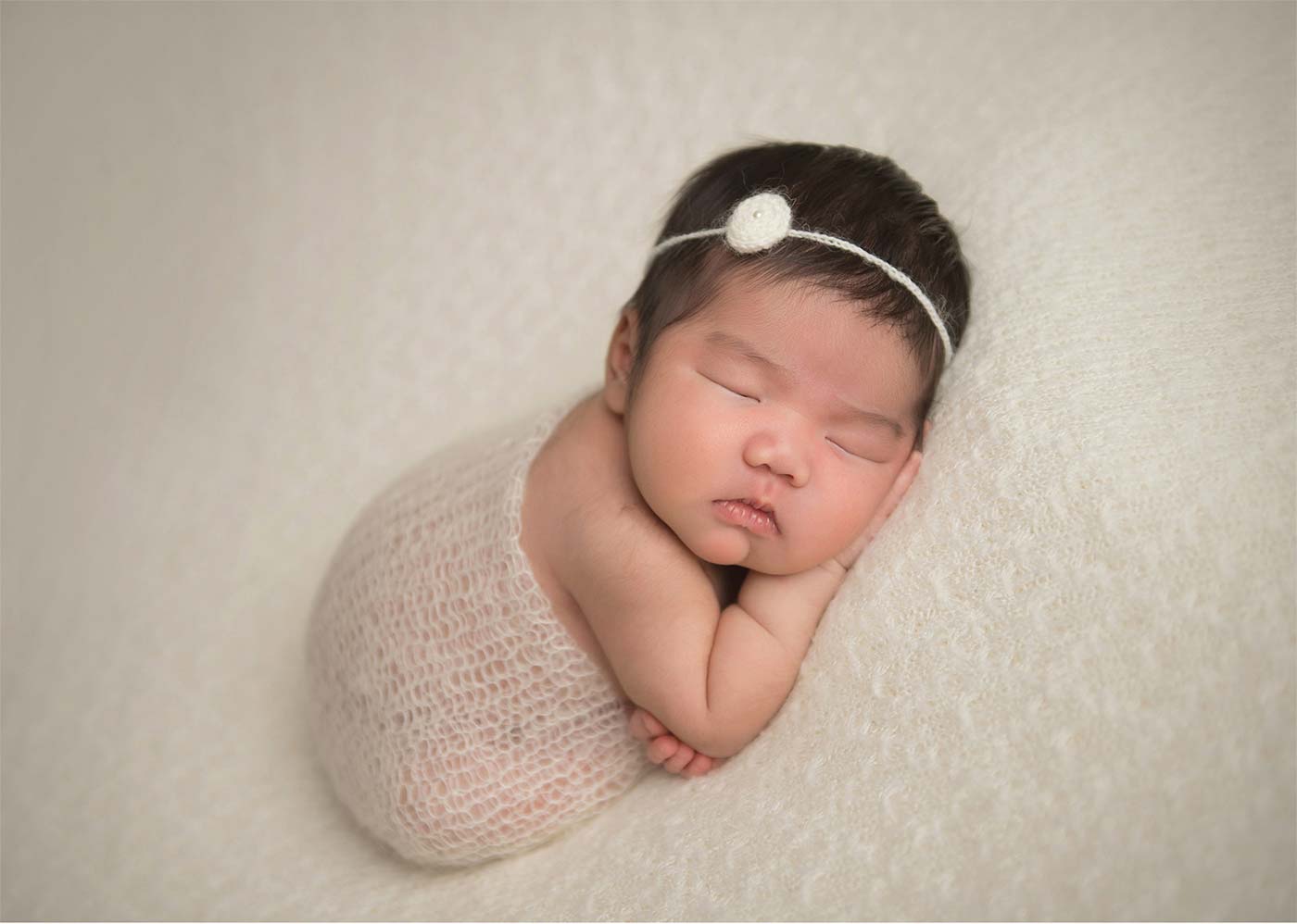 A newborn baby peacefully sleeps on their side, wrapped in a soft, cream-colored knitted blanket. The baby has a small headband adorned with a delicate flower, and they rest their head on their arms against a matching cream-colored background.