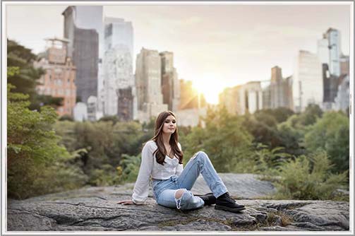 A stylish teen in ripped jeans posing for her senior portrait with the NYC skyline and sunset as the backdrop.